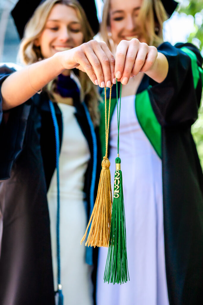 Two graduates hold colorful graduation tassels forward while smiling together.