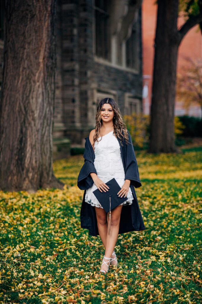 Full-length portrait of a graduate holding her cap while standing on a leaf-covered lawn.