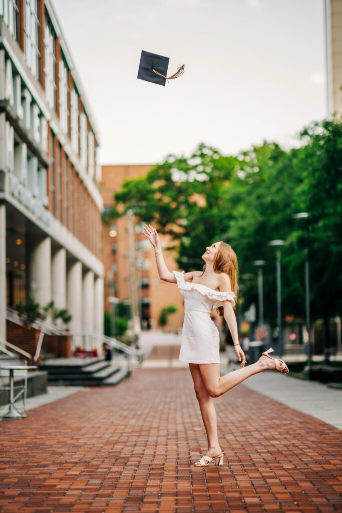 Graduate in a white dress tosses her cap into the air while standing on a brick campus walkway.