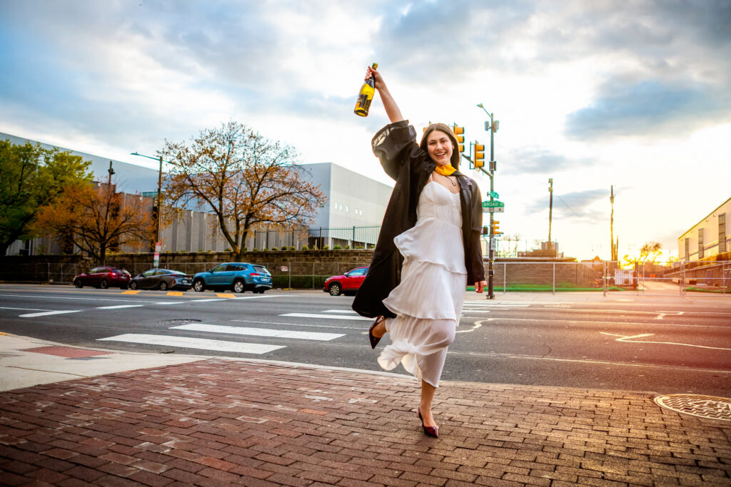 Graduate celebrates by jumping on Broad street in Philadelphia while holding a champagne bottle with the sun setting behind her.