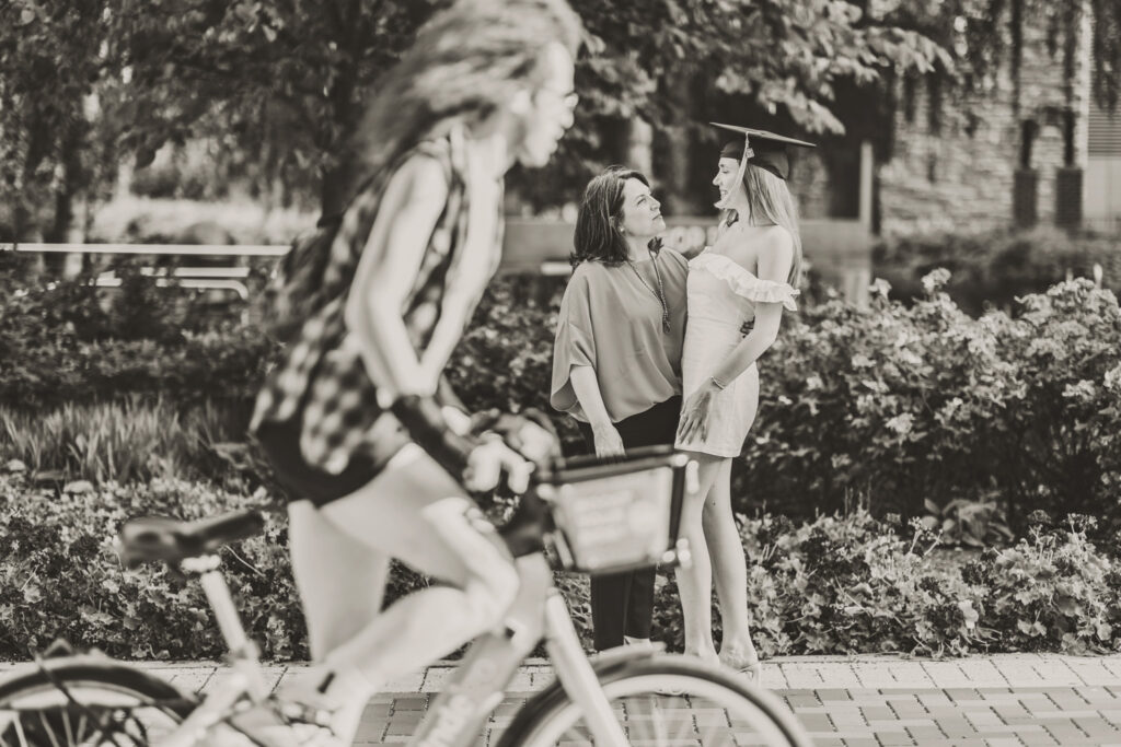 Black-and-white portrait of a graduate laughing with her mother during a graduation photoshoot while a bike and rider ride across the front of the frame.