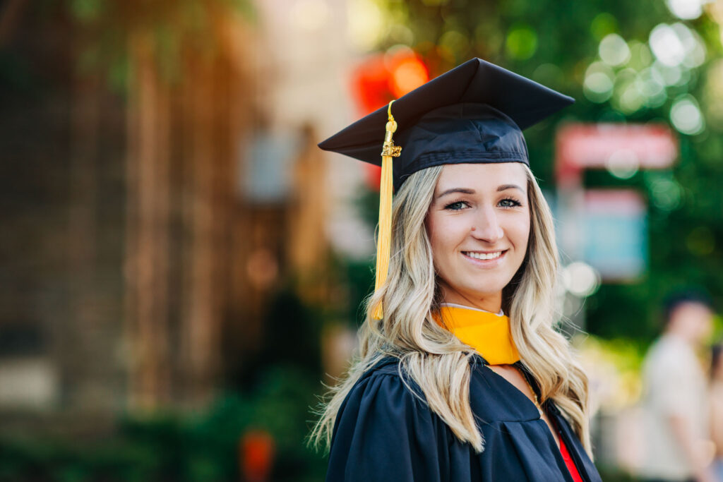 Graduate in cap and gown smiling at the camera during golden hour on campus with soft greenery in the background.
