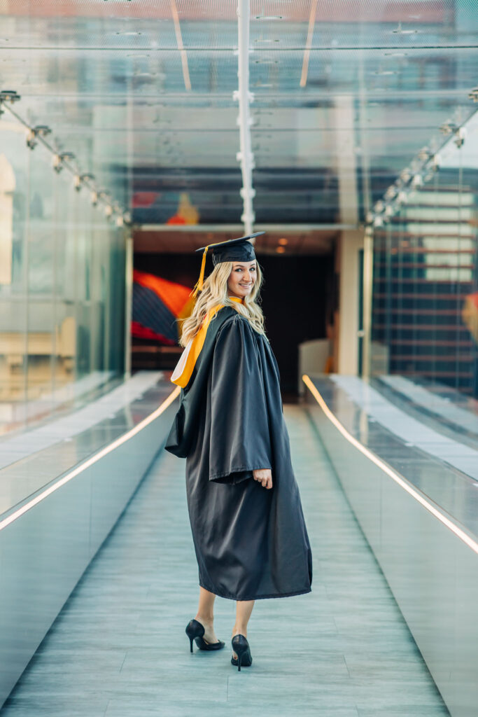 Graduate in a cap and gown looks back while walking across a modern glass campus bridge