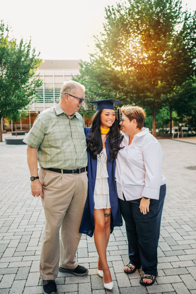 Graduate stands between family members, smiling together on a tree-lined campus walkway.