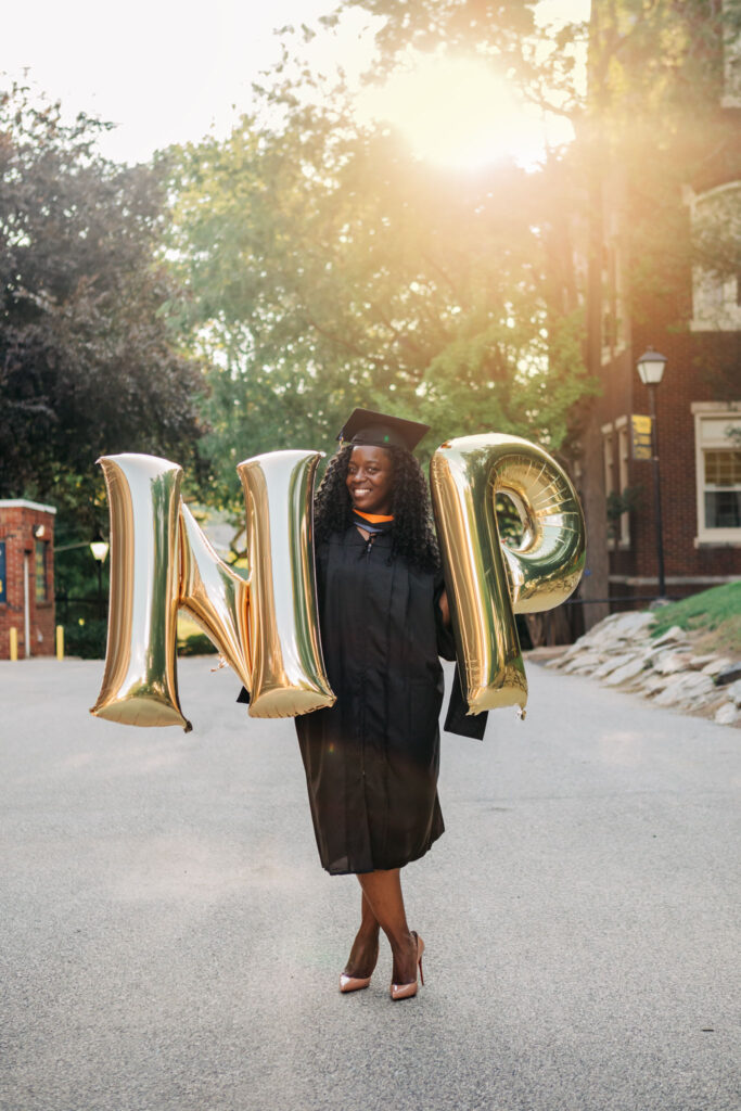 Graduate wearing a cap and gown holding large gold letter balloons outdoors during golden hour.
