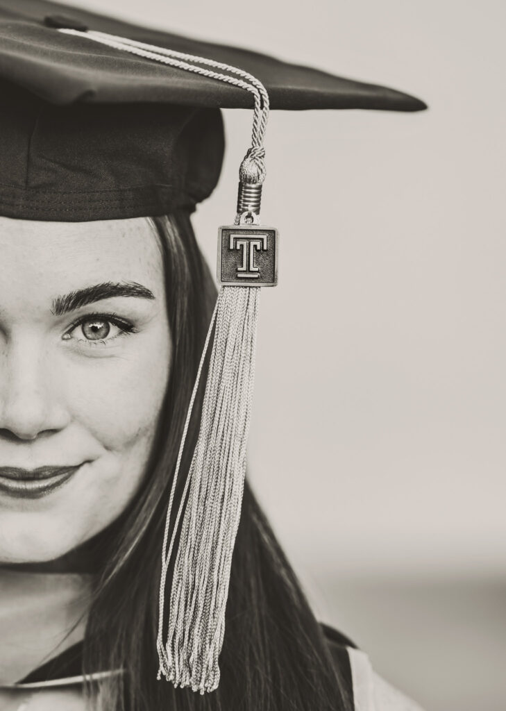 Black and white close-up of a female graduate's smiling face with a graduation cap wearing a Temple University tassel 