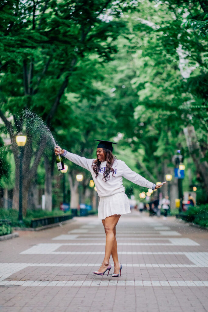 Graduate celebrating on a tree-lined campus path while spraying champagne, wearing a sweatshirt, skirt, and graduation cap.