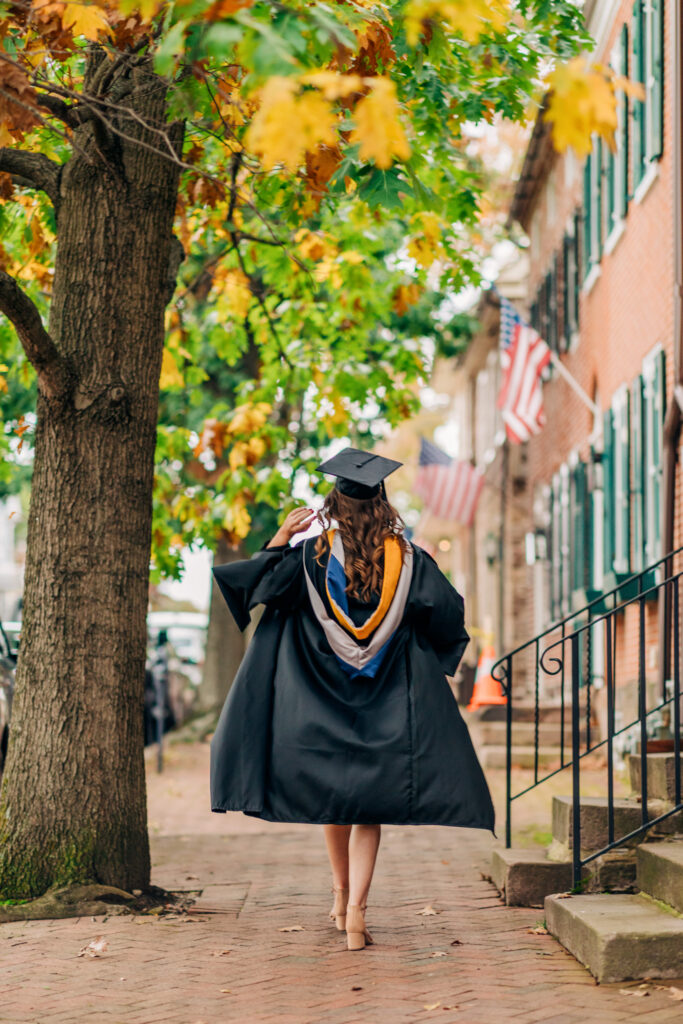 Graduate walks away down a tree-lined sidewalk with American flags in the background in a cap and gown during fall.