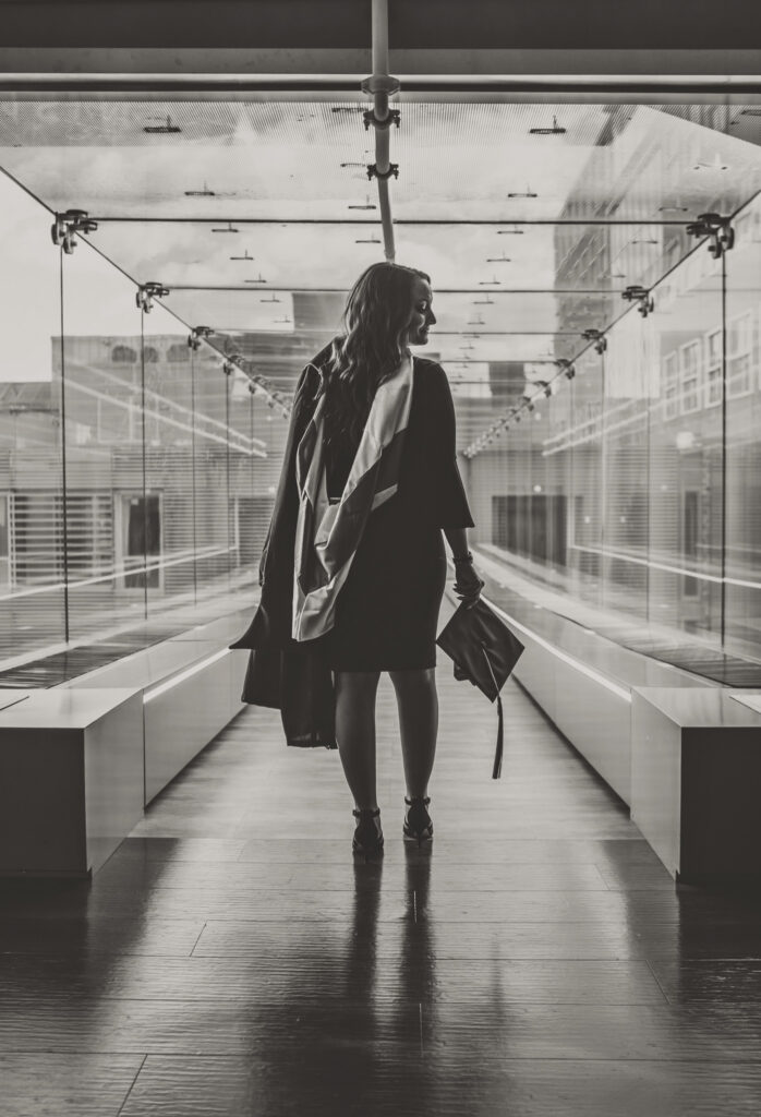 A black and white scene of a graduate wearing a gown and sash while holding a cap, standing in a quiet glass hallway with her body facing forward and her head turned to the side