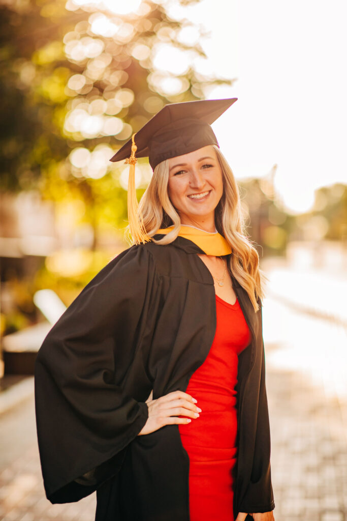 Female graduate with long blond hair and a red dress with black cap and gown smiling in cap and gown during a golden hour graduation photoshoot with warm natural light