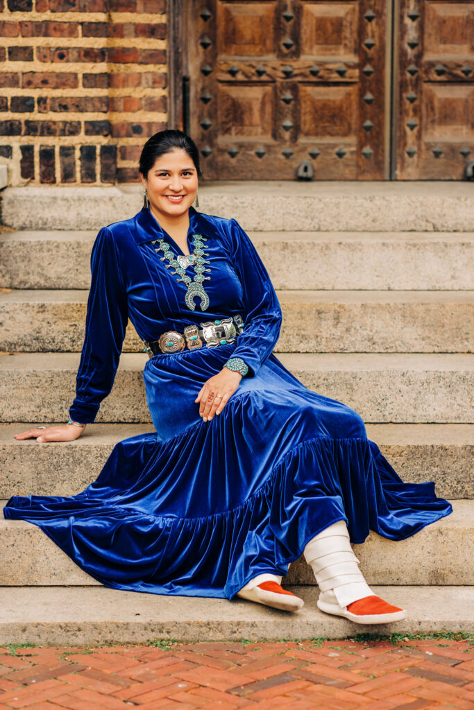 Graduate seated on stone steps wearing a blue velvet dress with turquoise jewelry and wrapped footwear, smiling at the camera.