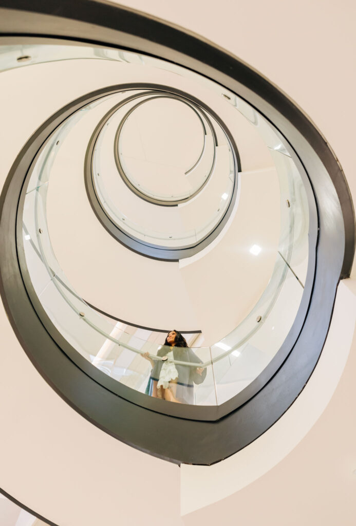 Graduate standing at the edge of a glass railing inside a circular spiral staircase, viewed from below
