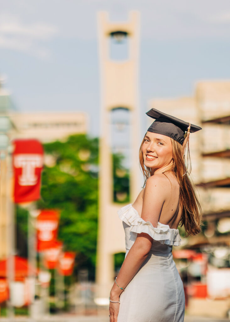 Graduate in an off-the-shoulder white dress and graduation cap, turning back toward the camera on a campus walkway.