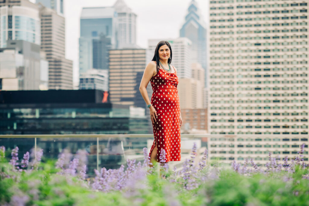 Graduate standing on a rooftop garden in a red patterned dress with the Philadelphia skyline in the background.
