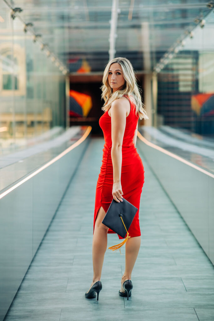 Graduate in a fitted red dress holding a graduation cap, standing on a modern glass walkway.