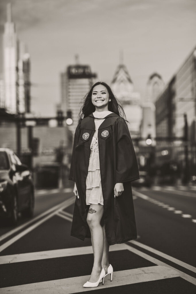 A black and white scene of a female graduate wearing a cap and gown and heels standing in a city street with the Philadelphia skyline softly blurred behind her