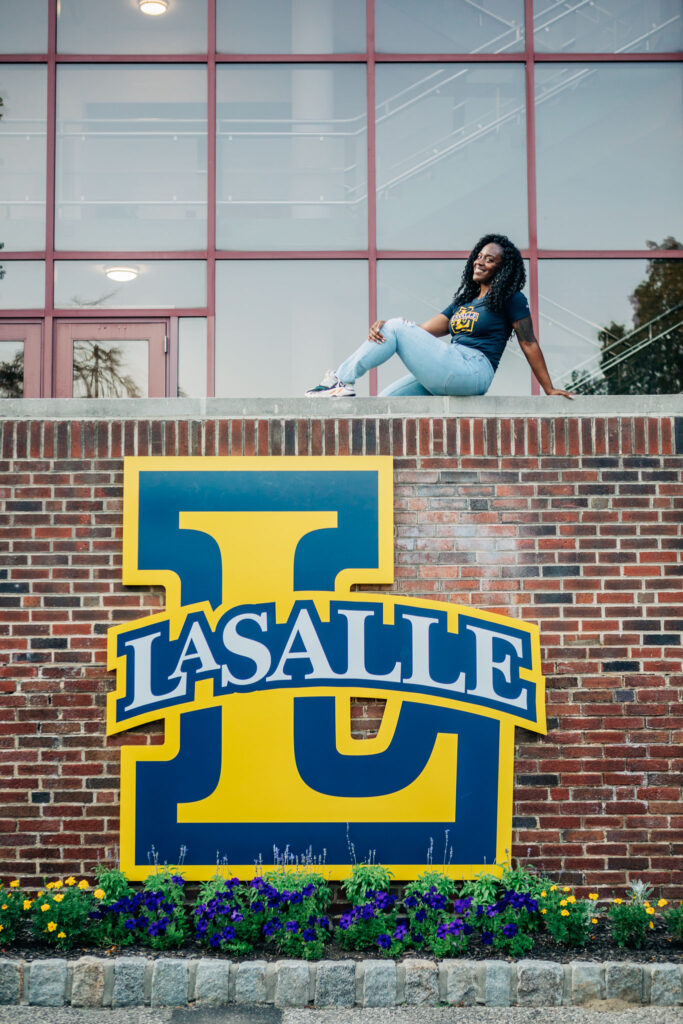 Graduate sitting on a brick ledge above the La Salle University sign on campus