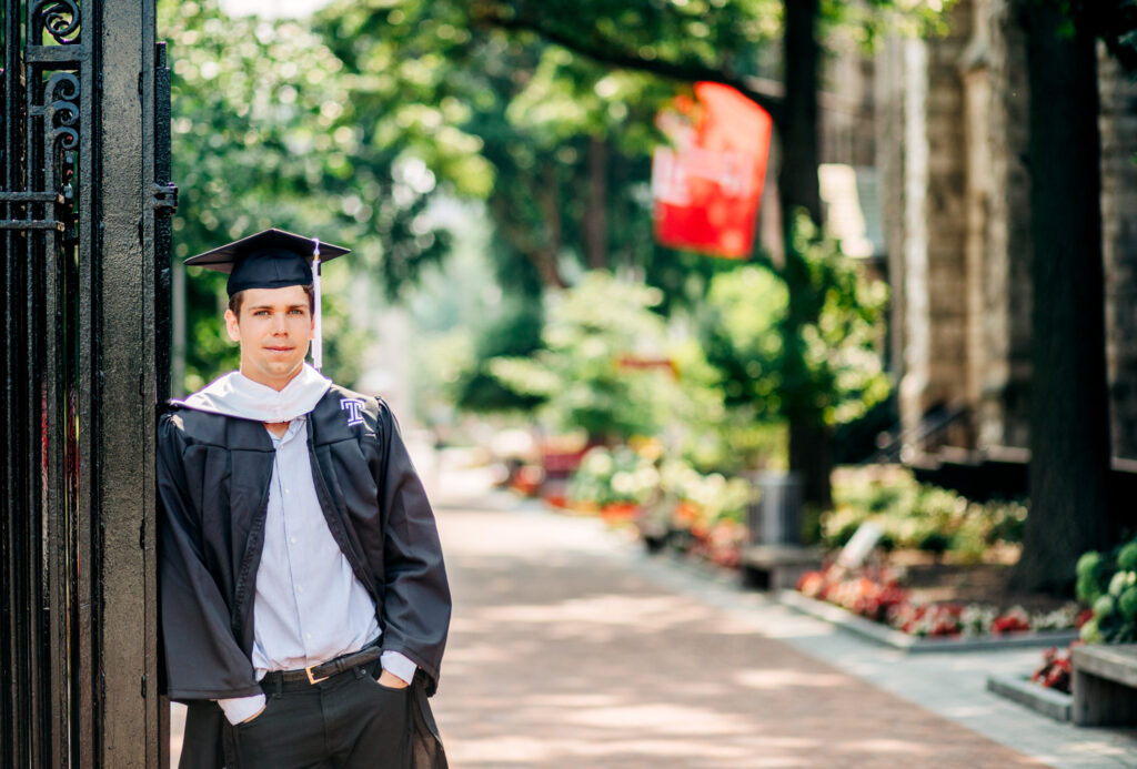 Male graduate leaning against a campus gate with hands in pockets, surrounded by greenery.