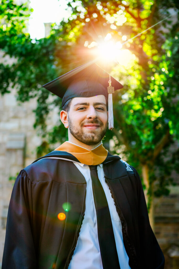 Male graduate wearing a cap and gown stands outdoors with sunlight filtering through trees behind him.