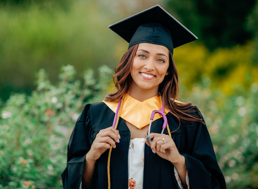 Nursing graduate wearing a cap and gown while holding a stethoscope in a garden setting.