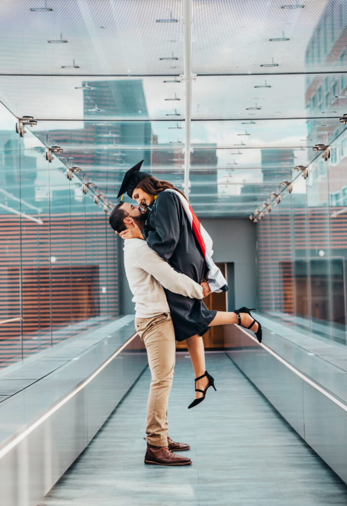 Graduate laughs as her partner lifts her in a glass walkway while celebrating graduation in a glass hallway.