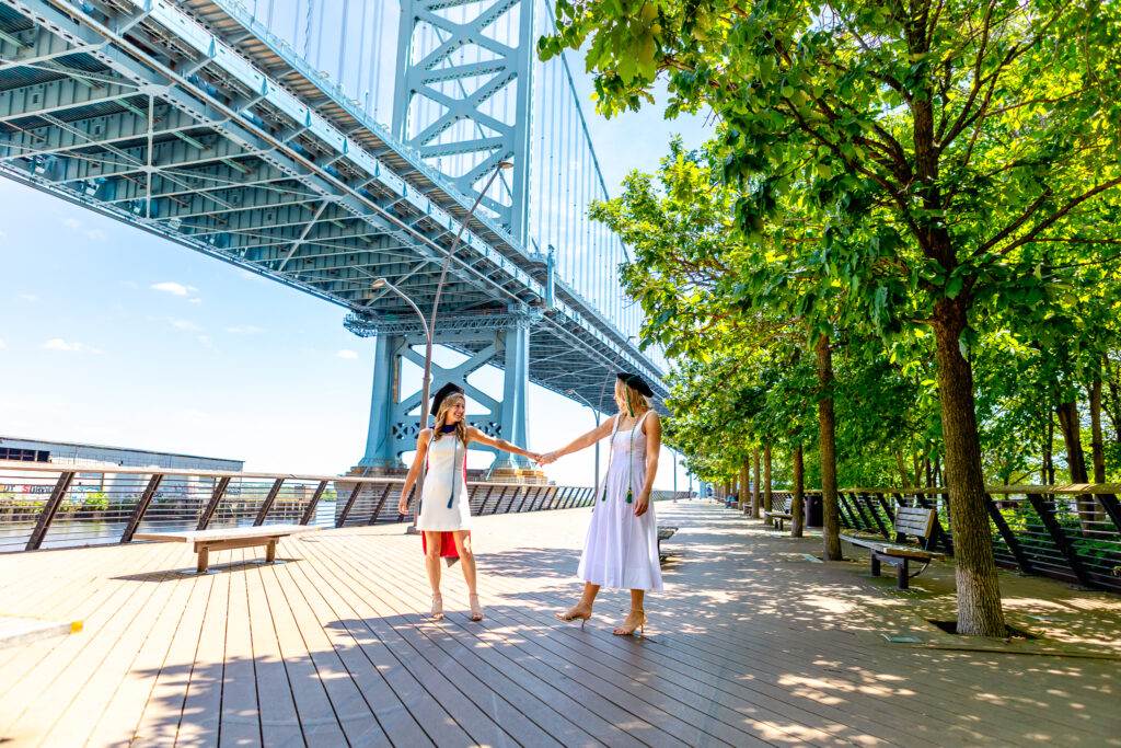Two graduates wearing white dresses and graduation stoles holding hands while walking along a riverside boardwalk beneath the Ben Franklin Bridge in Philadelphia