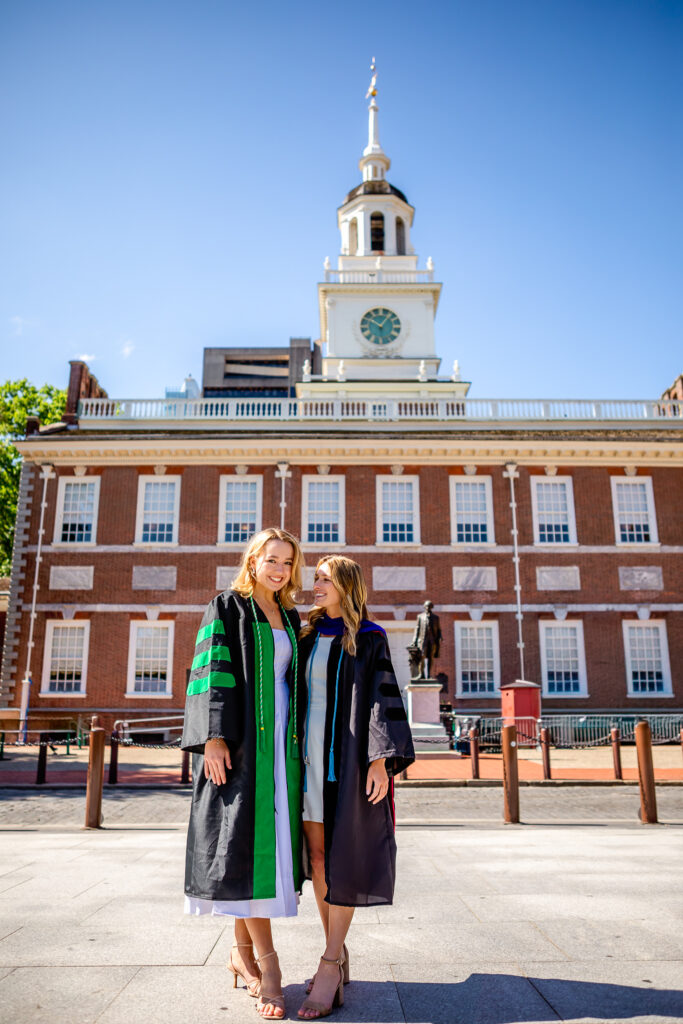 Two female college graduates posing in cap and gown during a graduation photoshoot in Philadelphia in front of Independence Hall