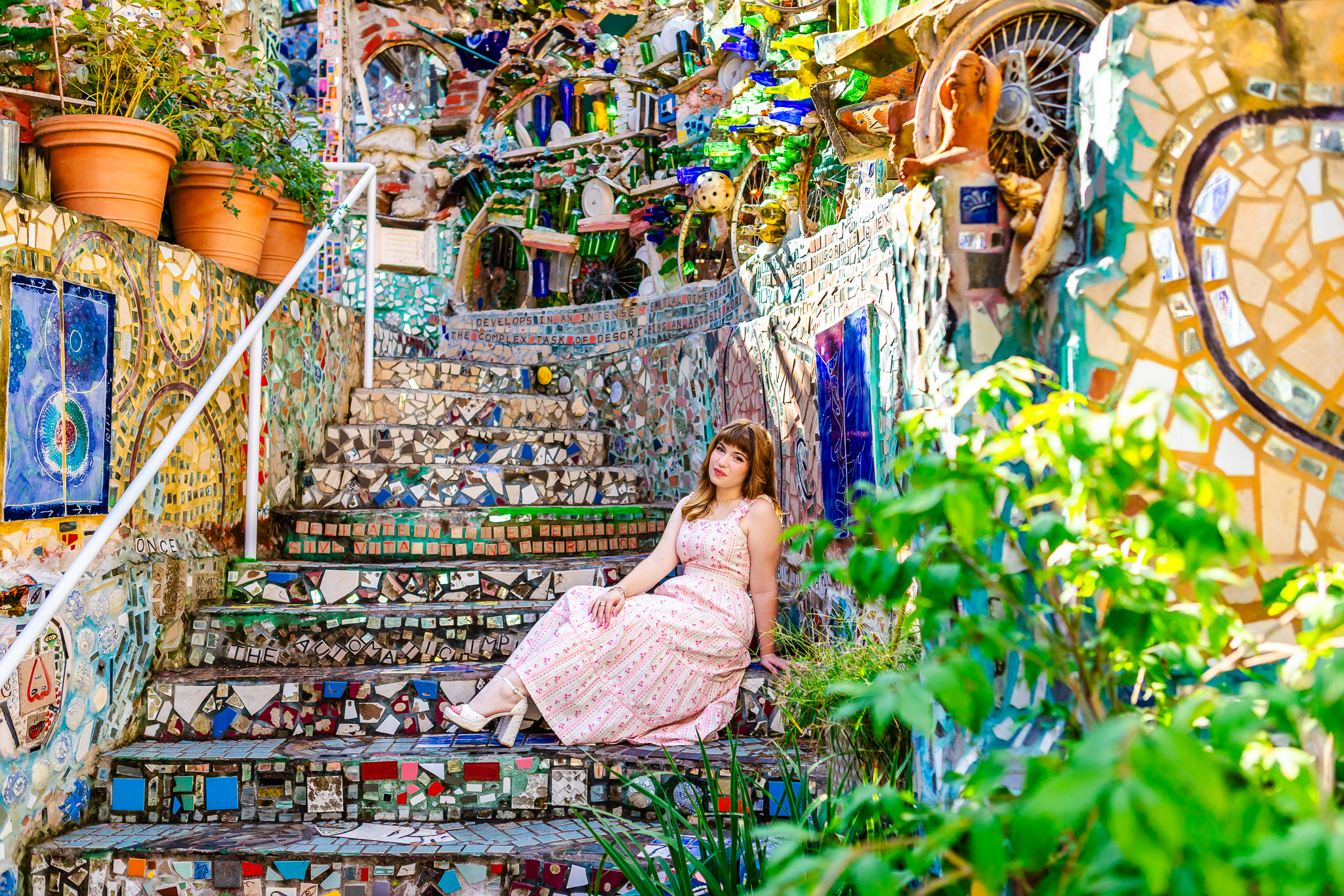 A high school senior in a long pink and white dress with red hair sits on the steps inside of Philadelphias Magic Garden surrounded by mosaic art.