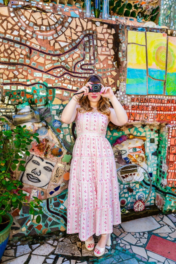 Full-length senior portrait of Lyla holding her camera while standing among vibrant mosaic artwork at Philadelphia Magic Gardens.