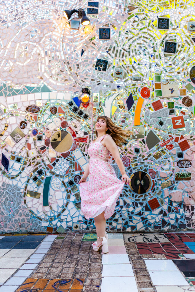 Senior portrait of Lyla twirling her dress in front of a vibrant mosaic wall. Her red hair is flowing behind her as she twirls.