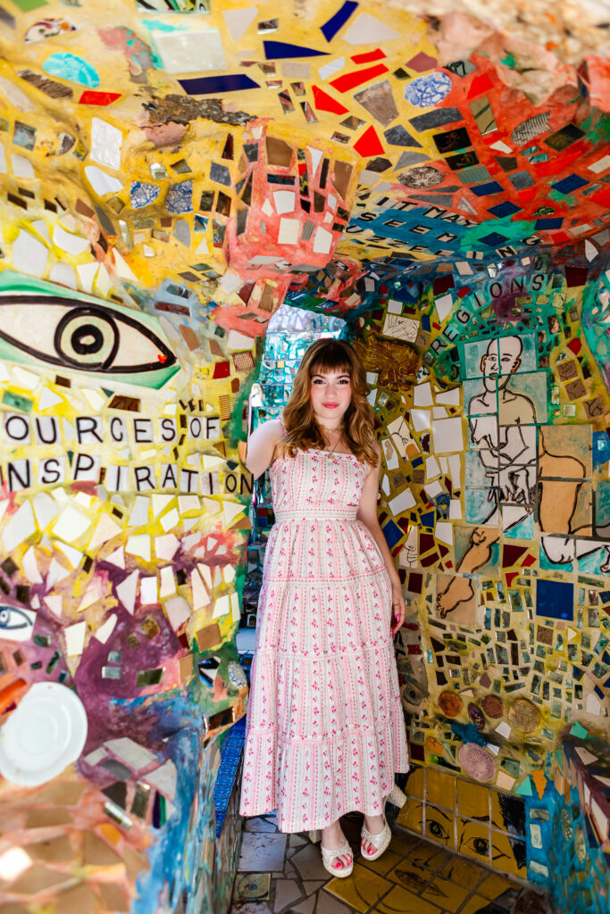 Full-body senior portrait of Lyla, wearing a white sundress with pink flowers and high heels, standing inside a colorful mosaic archway at the Magic Gardens