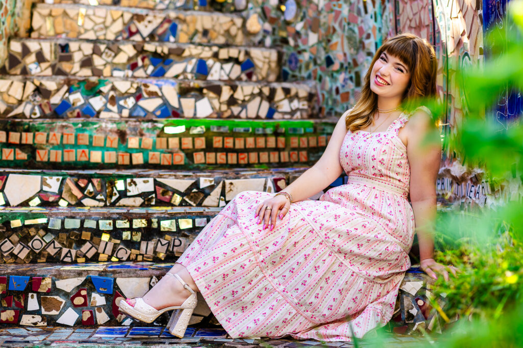 A female high school senior in a long pink sundress and long red hair with blunt bangs smiles at the camera with her head tilted back while sitting on the colorful mosaic steps inside the Philadelphia Magic Gardens.