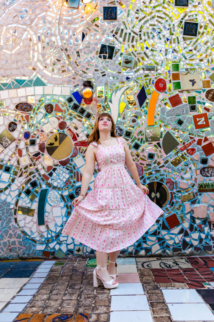 Senior portrait of Lyla looking upward and holding the bottom of her pink and white sundress out to the side while standing among detailed mosaic walls and artwork.