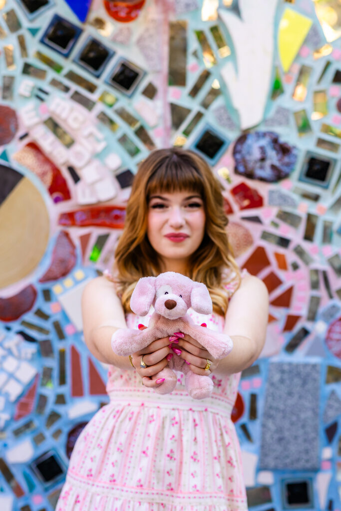 Senior portrait of Lyla extending her childhood stuffed animal toward the camera with mosaic artwork behind her.