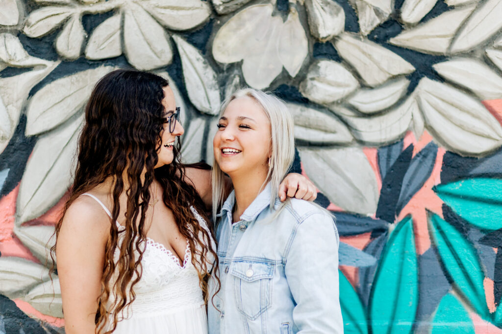 Two sisters stand together with their arms around each other in front of a colorful mural while laughing 