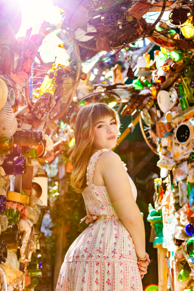 Senior portrait with warm backlight showing a student standing among mosaic walls and hanging glass artwork.