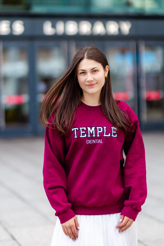 Temple University graduate wearing a Temple Dental sweatshirt and white skirt, standing outside a campus building.