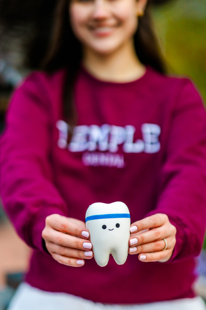Temple University dental graduate holding a smiling tooth prop in front of campus greenery.