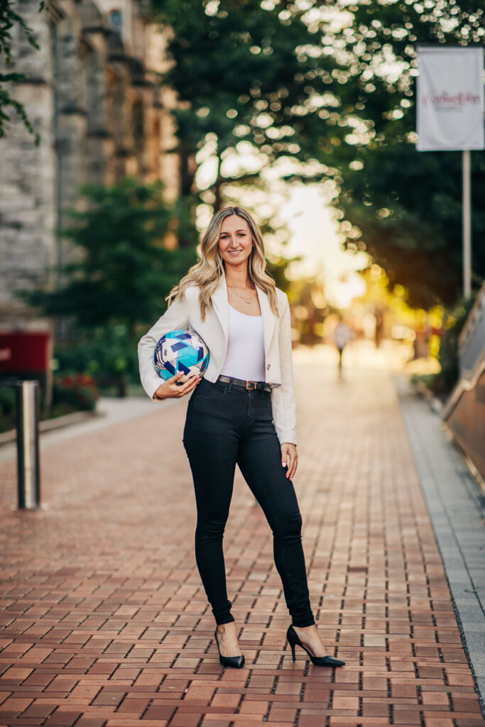 Temple University graduate with long blond hair wearing black pants and a short white coat holding a soccer ball on a brick walkway on campus with the sun setting behind her.