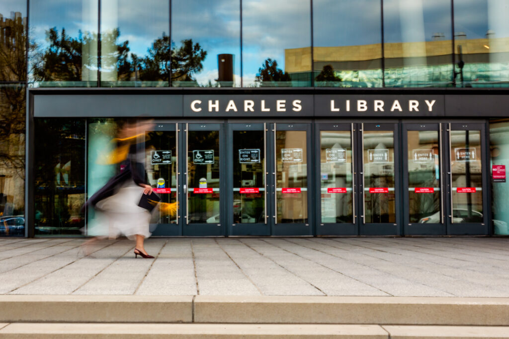 Graduate wearing a cap and gown walking in front of Charles Library at Temple University with intentional motion blur
