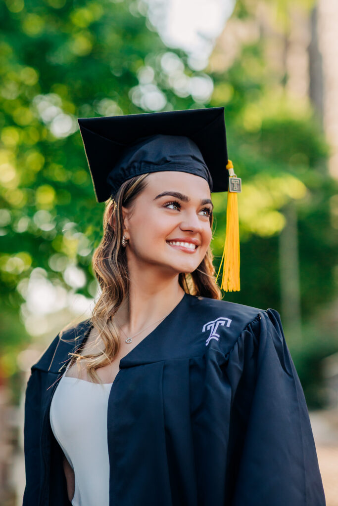 Graduate in cap and gown smiling while looking off camera on a sunlit campus path.