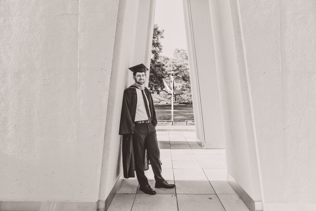 Black and white scene of a graduate in a cap and gown leaning against the inside of a bell tower with one hand in his pocket at Temple University