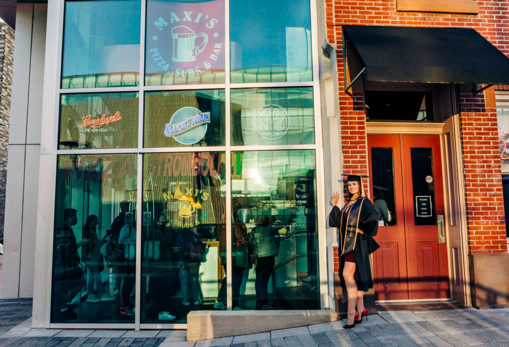 Graduate wearing a cap and gown and heels standing outside Maxi’s bar on Temple University's campus during a graduation portrait session