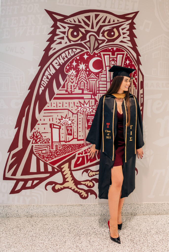 College graduate wearing a cap and gown and heels, standing in front of a Temple University owl mural