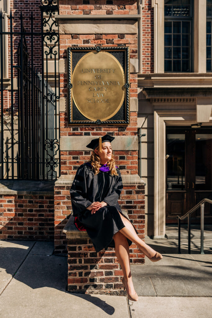 Graduate wearing academic regalia, sitting on a brick ledge beneath the University of Pennsylvania Carey Law School plaque with her legs crossed and her head turned to the side