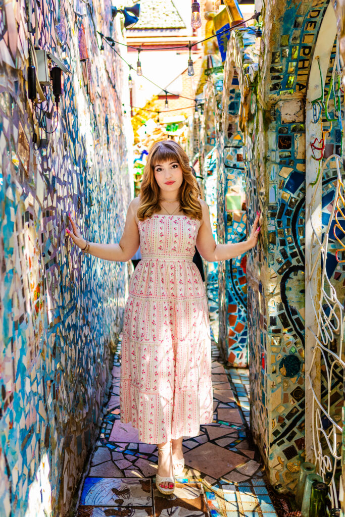 Urban senior portrait of a girl in a pink an white dress and long red hair with bangs standing between colorful mosaic walls with her arms outstretched and touching both walls.